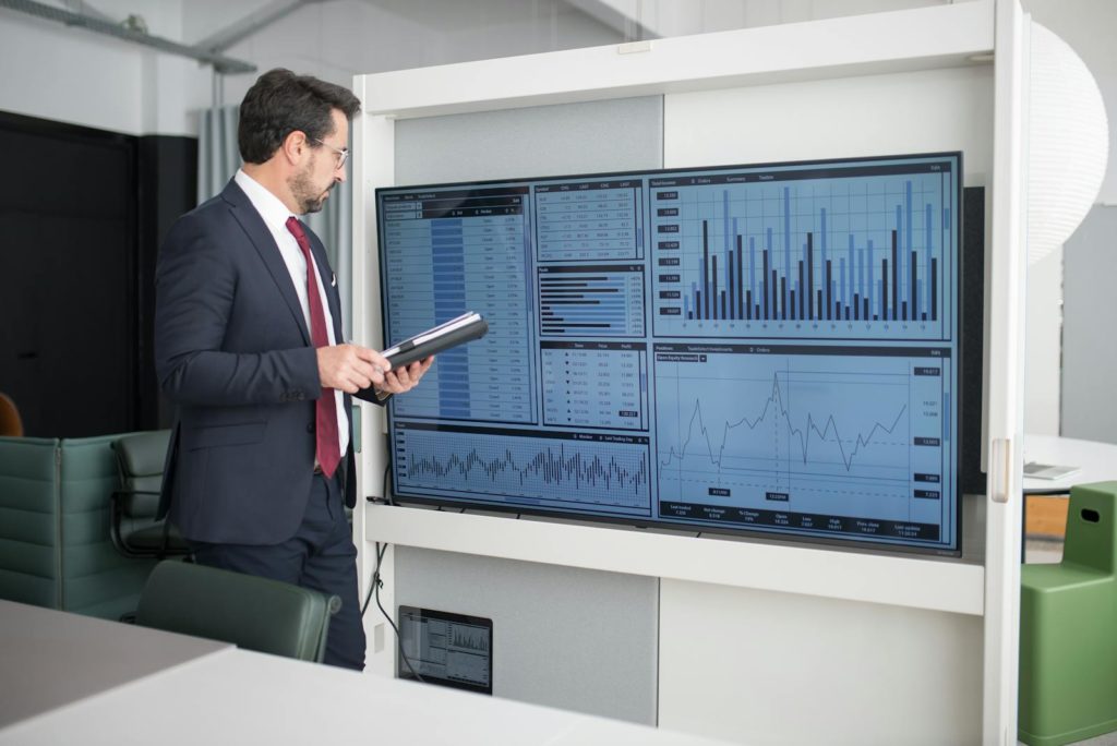 Un professionnel en costume examine des tableaux et des graphiques financiers sur un écran dans un bureau moderne.